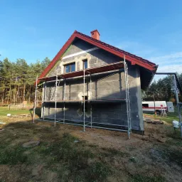 Dom jednorodzinny w trakcie ocieplania ścian zewnętrznych styropianem grafitowym, widoczne rusztowanie i fragment lasu w tle.