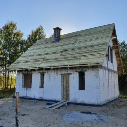 Surowy, biały dom z bloczków w trakcie budowy, z częściowo ukończoną drewnianą konstrukcją dachu i kominem, otoczony piaszczystym terenem budowy i drzewami.