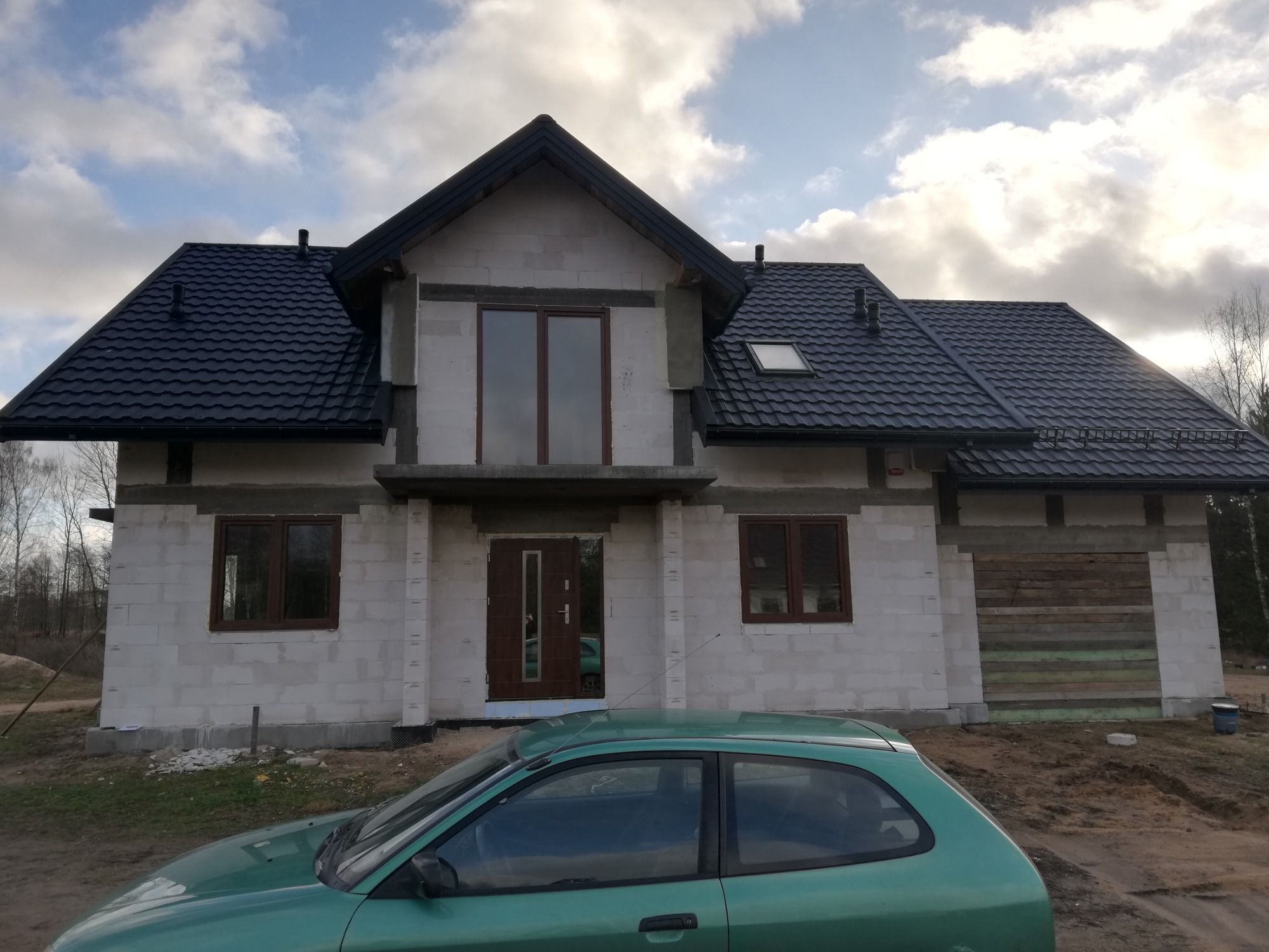 Surowy stan domu murowanego z bloczków, z czarną dachówką, widoczne okna i drzwi, częściowo zabezpieczone deskami, na pierwszym planie zielony samochód.