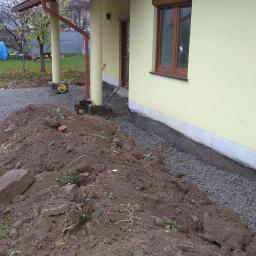 RKBUD - Wykonana izolacja fundamentów domu jednorodzinnego z widoczną membraną drenażową, obsypana żwirem i częściowo zasypana ziemią, z wykopem wzdłuż ściany budynku.