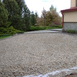 RKBUD - Teren przygotowany pod budowę, wysypany drobnym szarym żwirem, zabezpieczony geowłókniną, widoczne obrzeża i fragment budynku z kamienną elewacją.