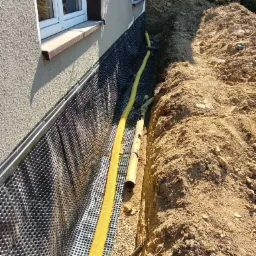 Odkryty fundament domu z folią kubełkową i żółtą rurą drenażową w wykopie wypełnionym żwirem, ziemia w tle.