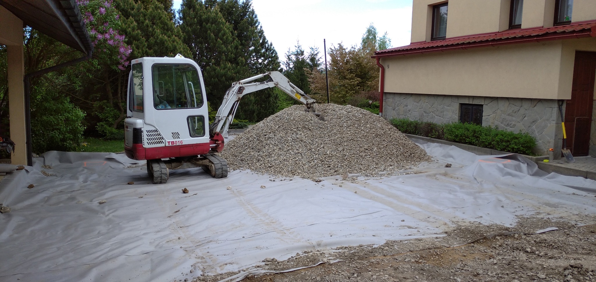 Mini koparka TB016 rozładowuje stertę żwiru na rozłożoną geowłókninę, przygotowanie terenu pod budowę domu z kamienną podmurówką.