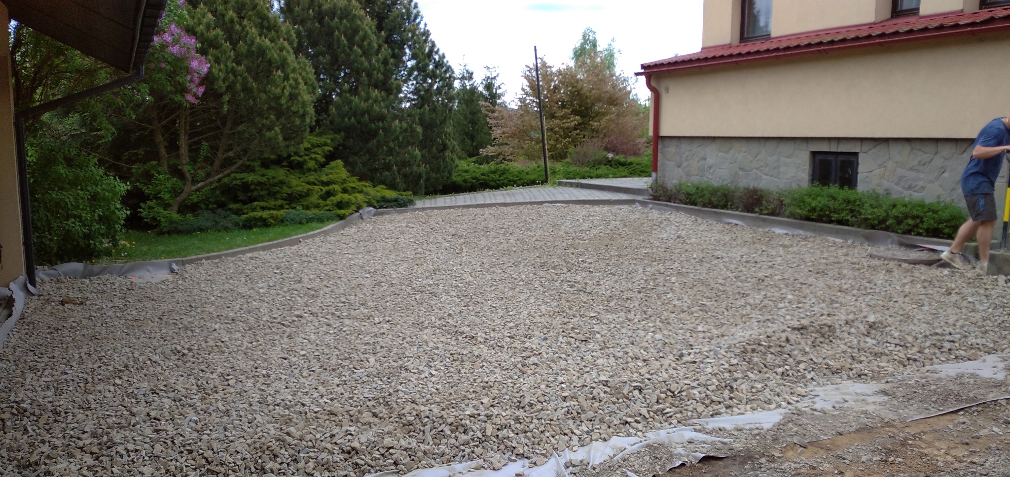 Teren przygotowany pod budowę, wysypany drobnym szarym żwirem, zabezpieczony geowłókniną, widoczne obrzeża i fragment budynku z kamienną elewacją.