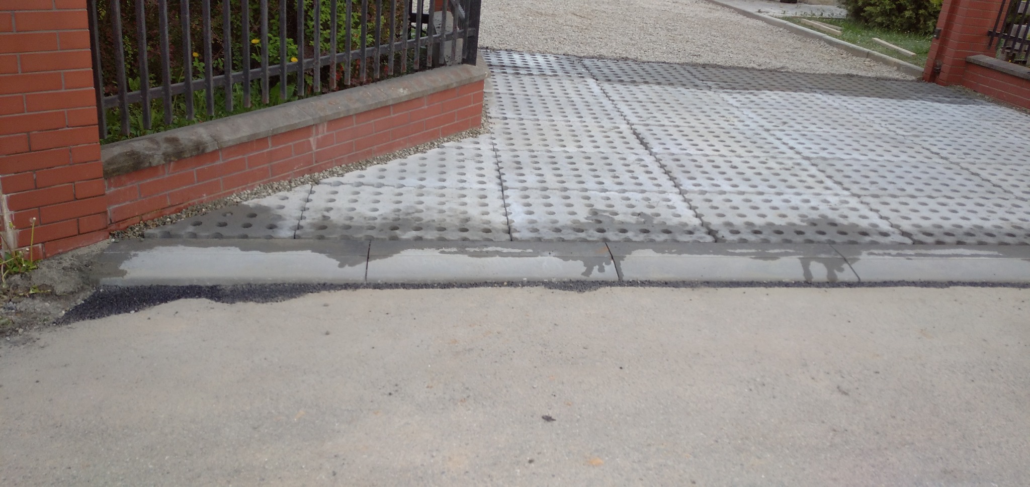 Ułożona nawierzchnia z ażurowych betonowych płyt obrzeżona betonowym krawężnikiem, widok z poziomu gruntu, częściowo widoczny ceglany murek z metalowym ogrodzeniem.