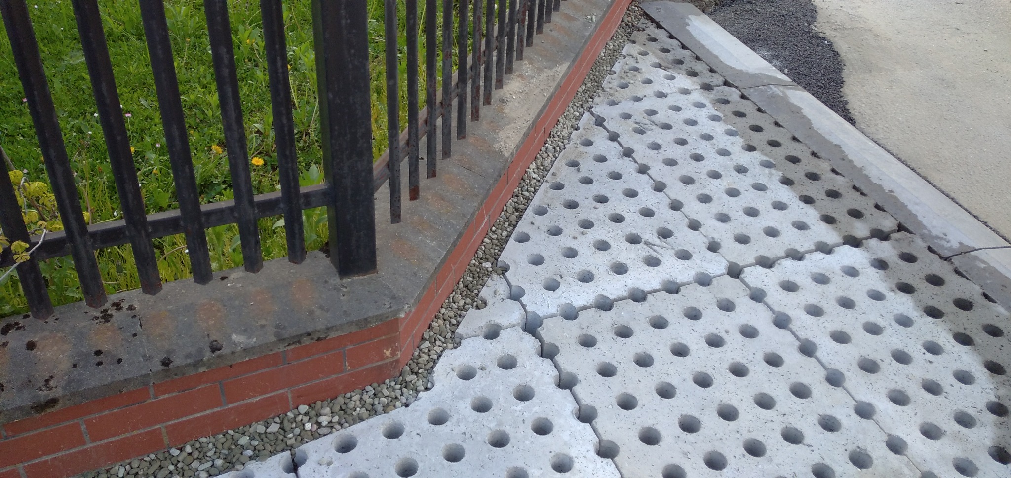 Szare, betonowe płyty ażurowe z okrągłymi otworami, ułożone na podłożu z drobnych kamieni, przylegające do ceglanego murku z metalowym ogrodzeniem. Widoczne pęknięcia między niektórymi płytami.