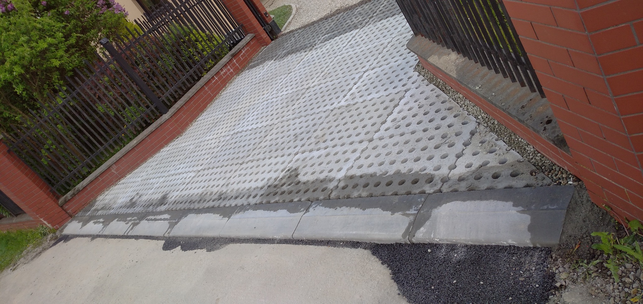 Ułożona, szara, ażurowa kostka na podjeździe, obrzeża z betonowych krawężników, widoczny fragment metalowego ogrodzenia i ceglanej bramy.