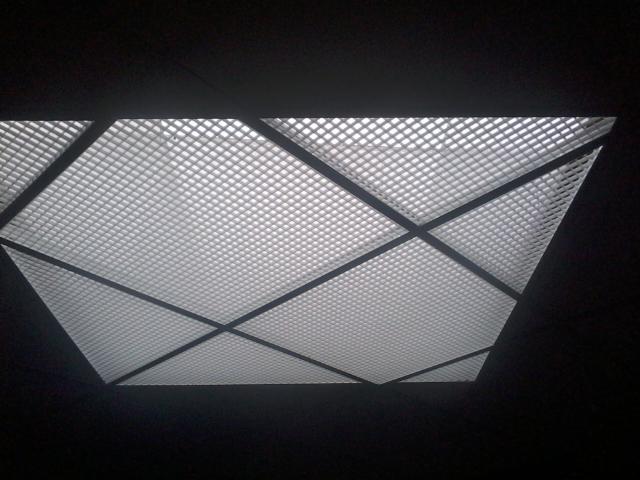 Kaseton sufitowy z kratką, widoczny z dołu, oświetlony od tyłu. Ciemne tło podkreśla geometryczny wzór i jasność paneli. Metalowa konstrukcja tworzy linie dzielące powierzchnię.