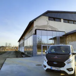 Biały samoch&oacute;d dostawczy Ford z logo firmy montującej stolarkę aluminiową zaparkowany przed budynkiem z nową szklaną fasadą z ramami okiennymi, widoczne odbicia w szybach.