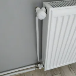 Biały, żebrowany grzejnik z termostatem na tle szarej ściany i paneli podłogowych imitujących drewno.