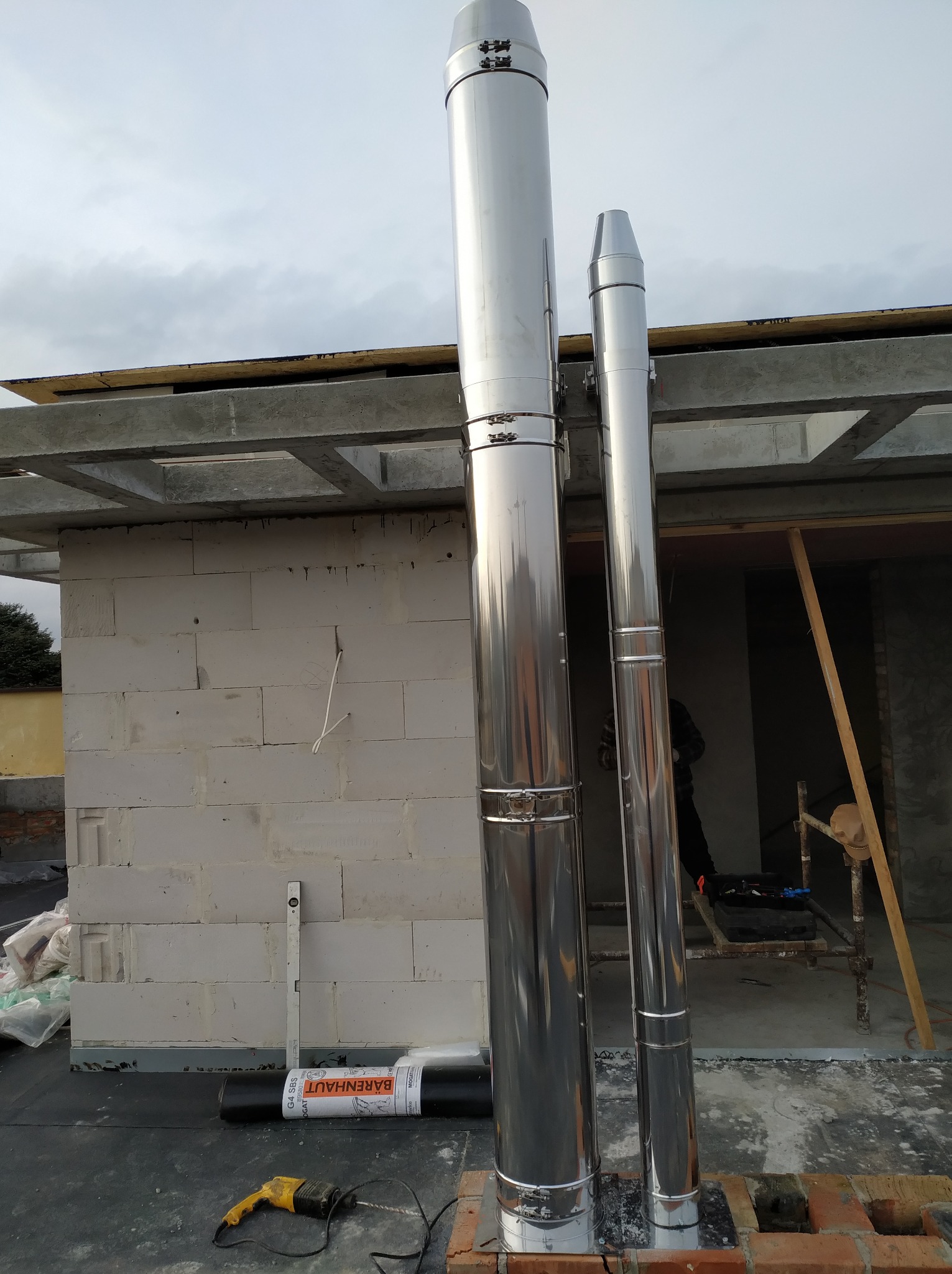 Dwa błyszczące, stalowe kominy zamontowane na dachu budynku w trakcie budowy, widoczny poziomica, zwój papy i wiertarka.