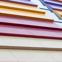 Fragment elewacji budynku z poziomymi panelami w kolorach: żółtym, czerwonym, fioletowym i kremowym, z widoczną metalową konstrukcją wsporczą.