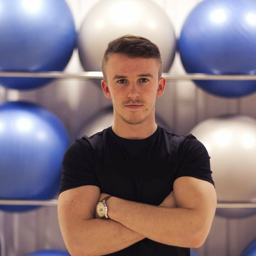 Portret trenera personalnego z założonymi rękami, w tle piłki do ćwiczeń w kolorach niebieskim i srebrnym, z widocznym zegarkiem na nadgarstku.