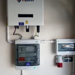 Biały falownik Solplanet i rozdzielnice PV-DC oraz PV-AC zamontowane na ścianie, widoczne przewody i naklejki ostrzegawcze.