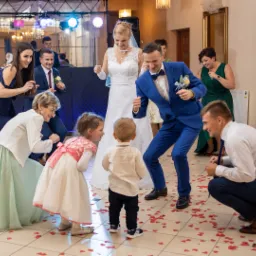 Para młoda w tańcu z gośćmi weselnymi, w tym z dziećmi, na parkiecie posypanym czerwonymi serduszkami, oświetlonym reflektorami.