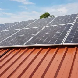 Panele fotowoltaiczne zamontowane na dachu z blachy trapezowej w kolorze ceglastym, widoczne mocowania i przewody, błękitne niebo w tle.