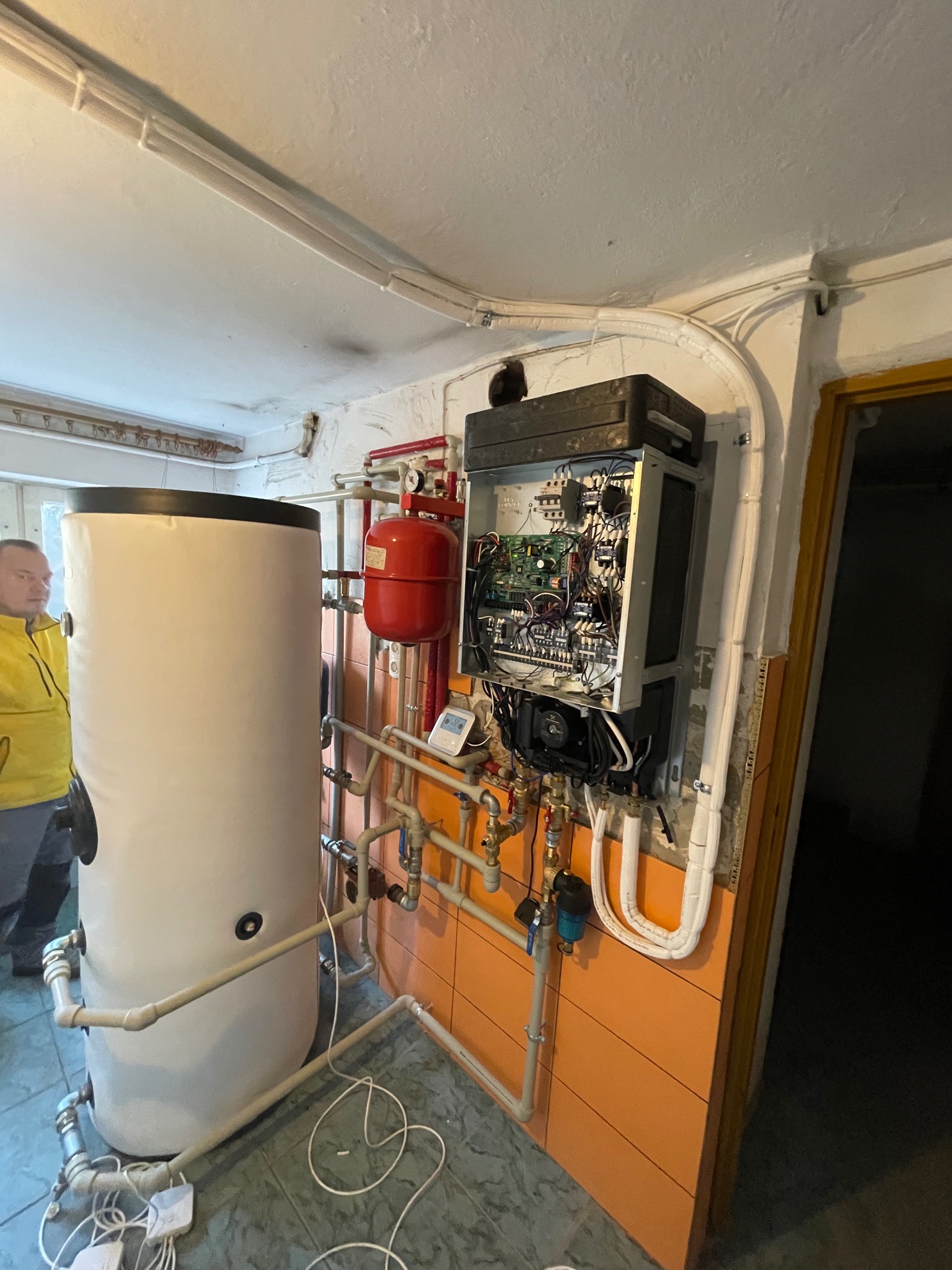 Otwarta obudowa pompy ciepła z widocznymi podzespołami elektronicznymi i hydraulicznymi, zbiornik akumulacyjny, instalacja rurowa w piwnicy z pomarańczowymi płytkami, z fragmentem osoby w żółtej...