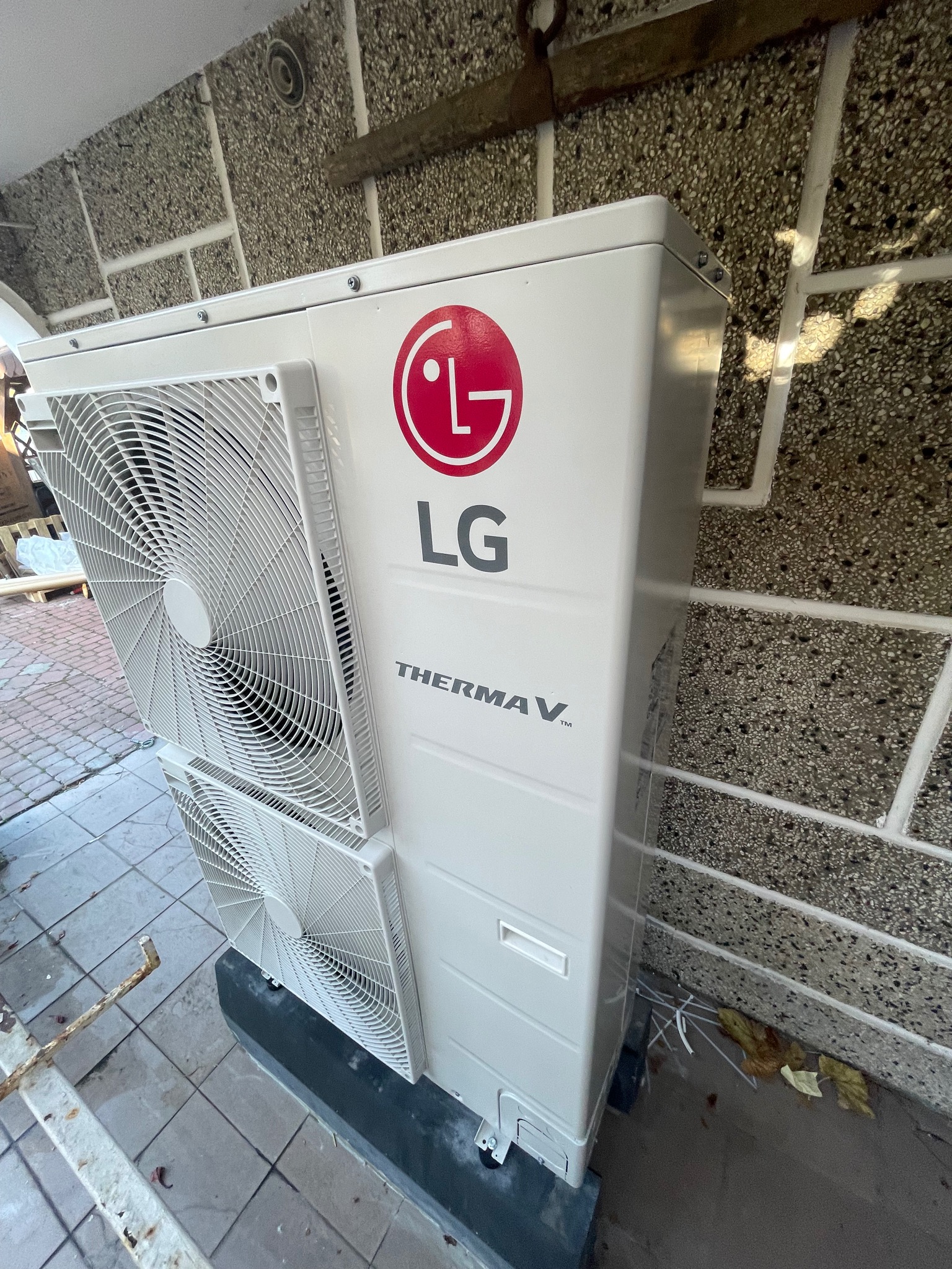 Zewnętrzna jednostka pompy ciepła LG Therma V z dwoma wentylatorami, zamontowana przy ścianie z fakturą kamieni ozdobnych i widocznymi przewodami na posadzce.