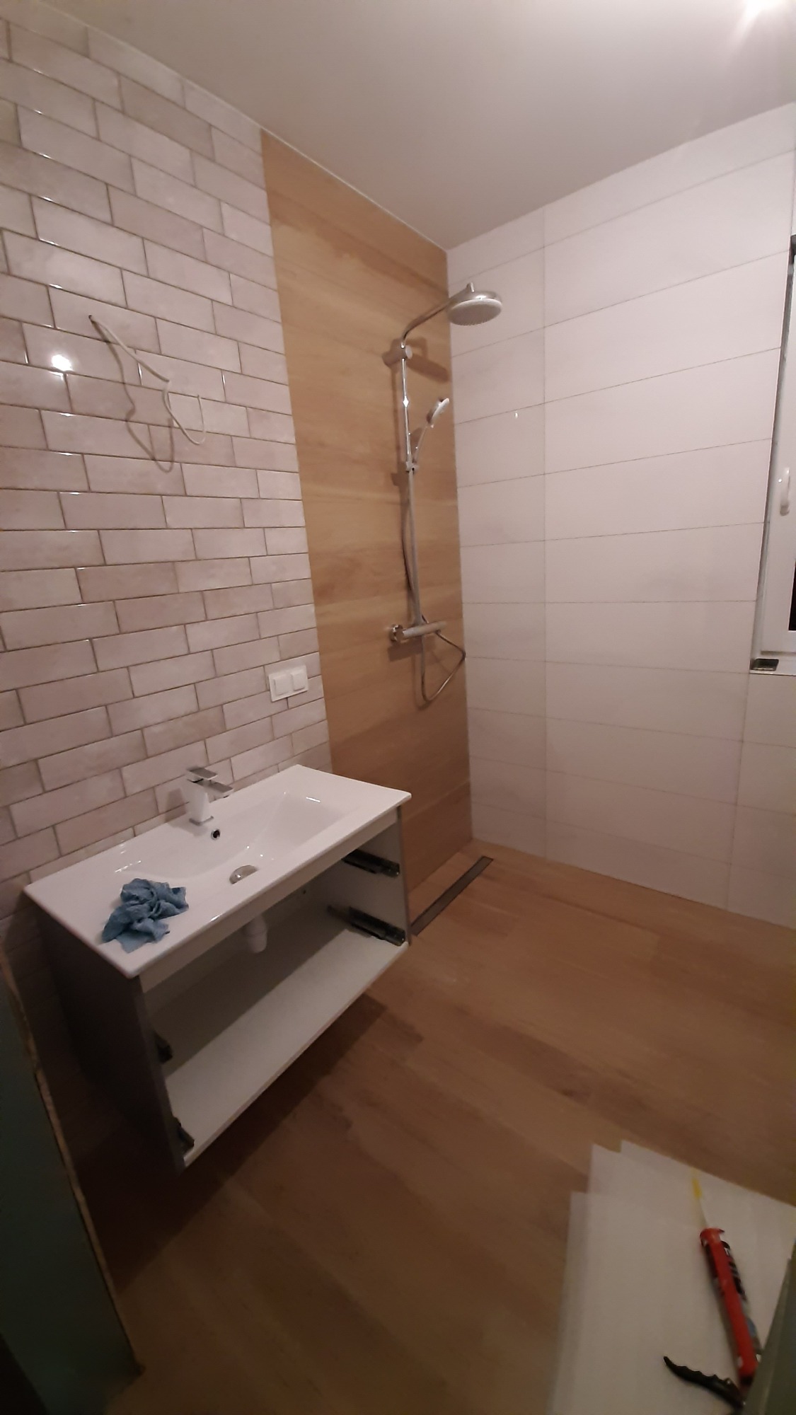 Ukończona łazienka z umywalką nablatową, baterią i ręcznikiem, kabina prysznicowa bez drzwi, ściany wyłożone białymi i drewnopodobnymi płytkami, na podłodze drewnopodobne płytki, widoczne narzędzia...