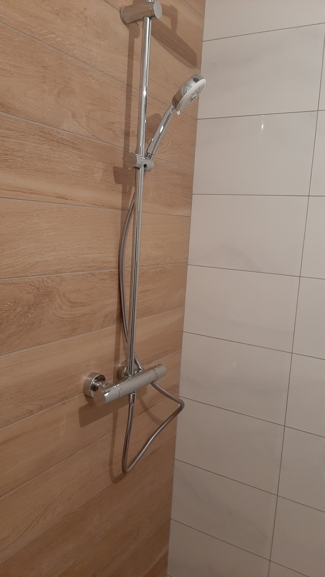 Chromowany zestaw prysznicowy z termostatem, zamontowany na ścianie wyłożonej płytkami imitującymi drewno, obok ściany z białych, prostokątnych płytek.
