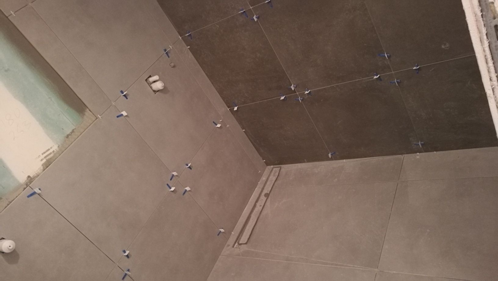 Układanie szarych płytek w łazience z użyciem krzyżyków dystansowych, widoczny odpływ liniowy i częściowo przygotowana ściana do dalszych prac wykończeniowych.