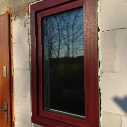 Ciemnoczerwone okno z widoczną pianką montażową po obwodzie, osadzone w ścianie z betonu komórkowego, z refleksami drzew na szybie i fragmentem drewnianych drzwi po lewej stronie.