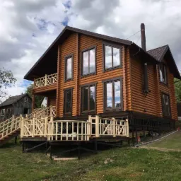 Dwukondygnacyjny dom z bali na fundamencie z drewnianym gankiem i schodami, widok z dołu z boku, na drugim planie stara drewniana szopa.