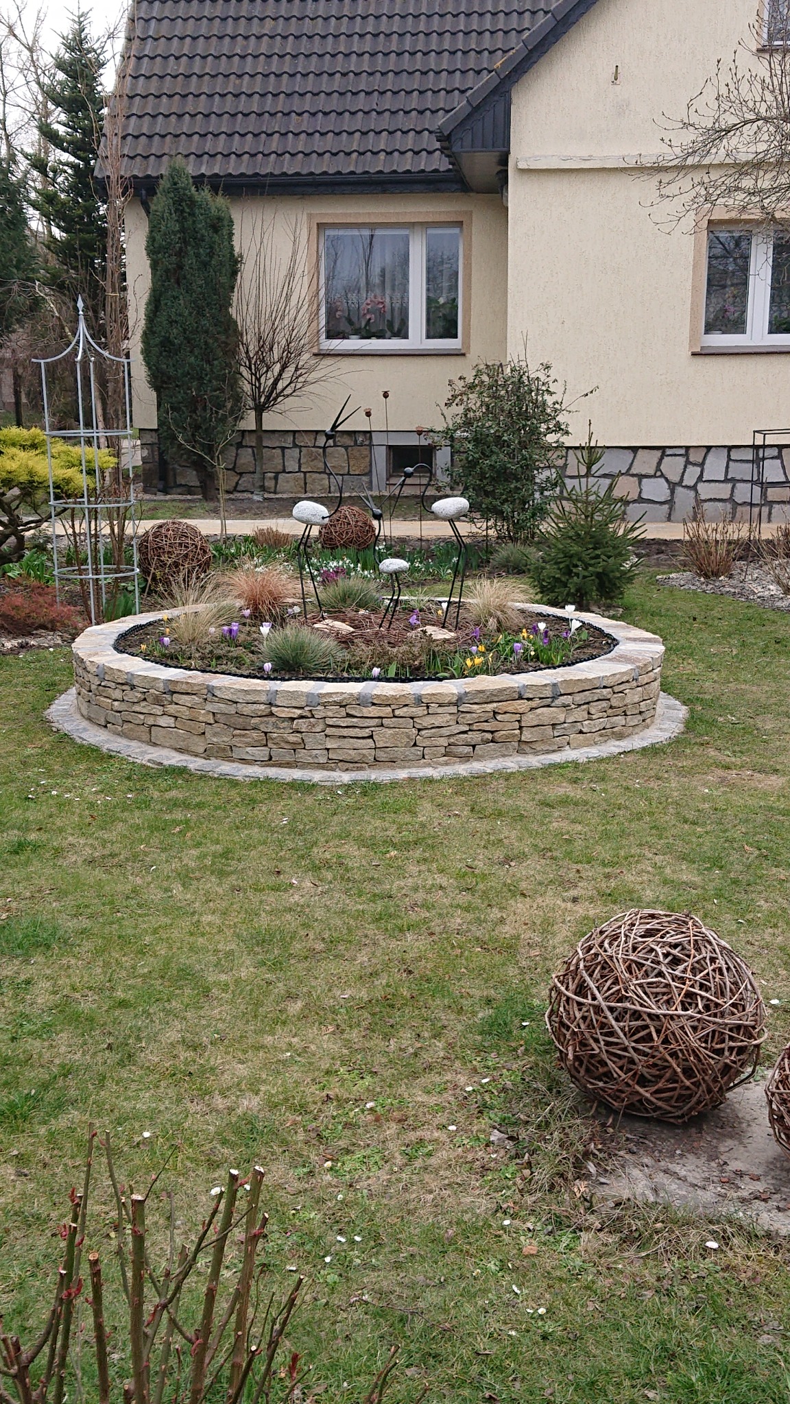 Okrągła rabata z kamienia polnego w ogrodzie przydomowym, ozdobiona metalowymi figurkami ptaków i kulami z wikliny.