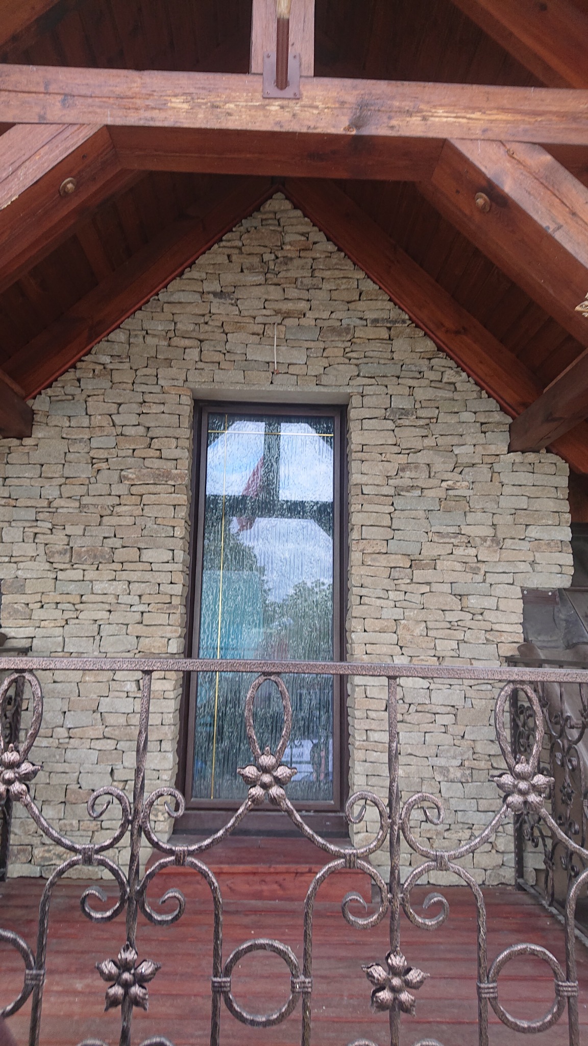 Kamienna elewacja domu z oknem, drewnianą konstrukcją dachu i ozdobną, metalową balustradą z motywem kwiatowym.