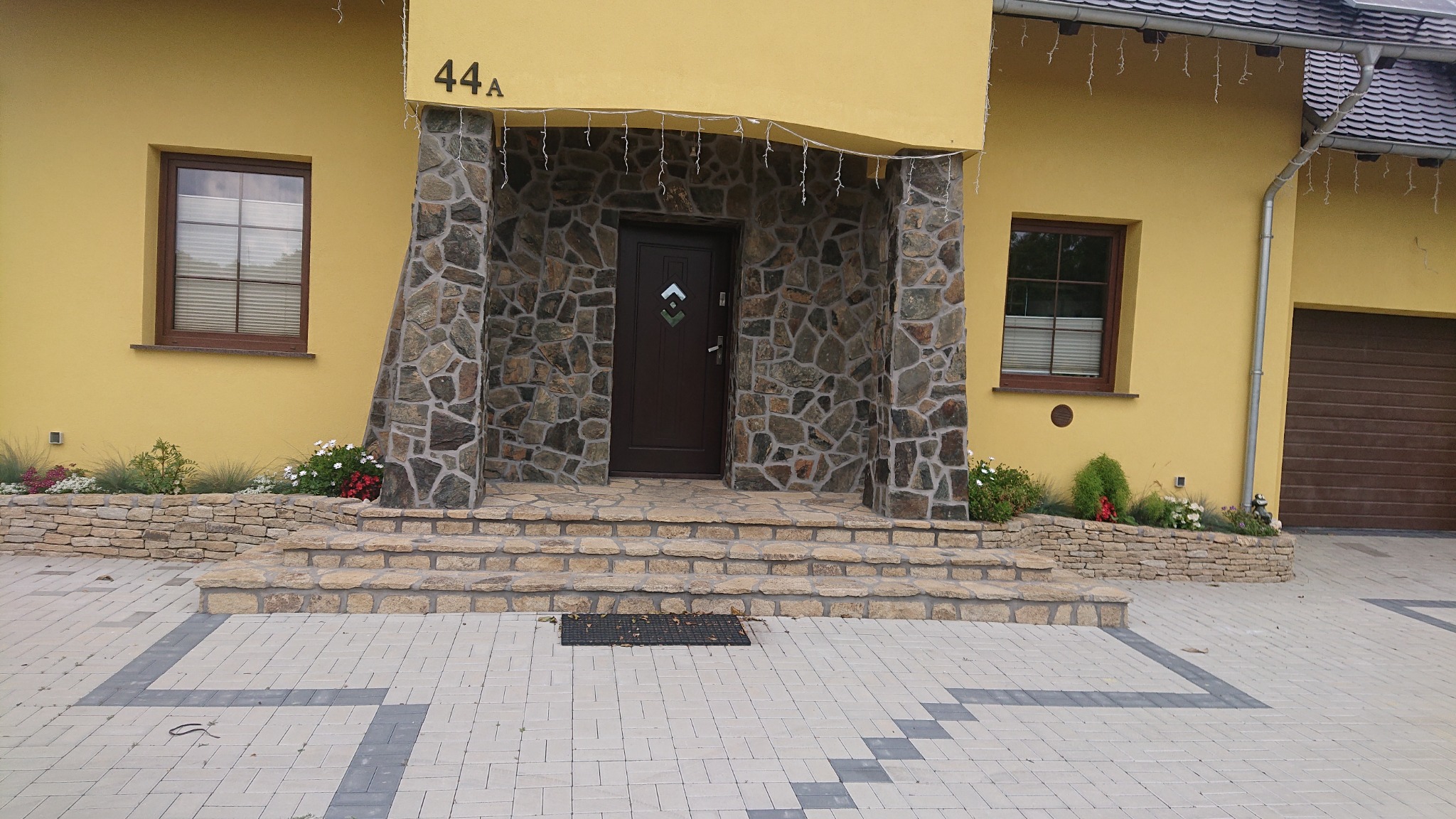 Wejście do domu z brązowymi drzwiami, kamienną kolumną i schodami, widok na żółtą fasadę z oknami i garażem, kostka brukowa na podjeździe.