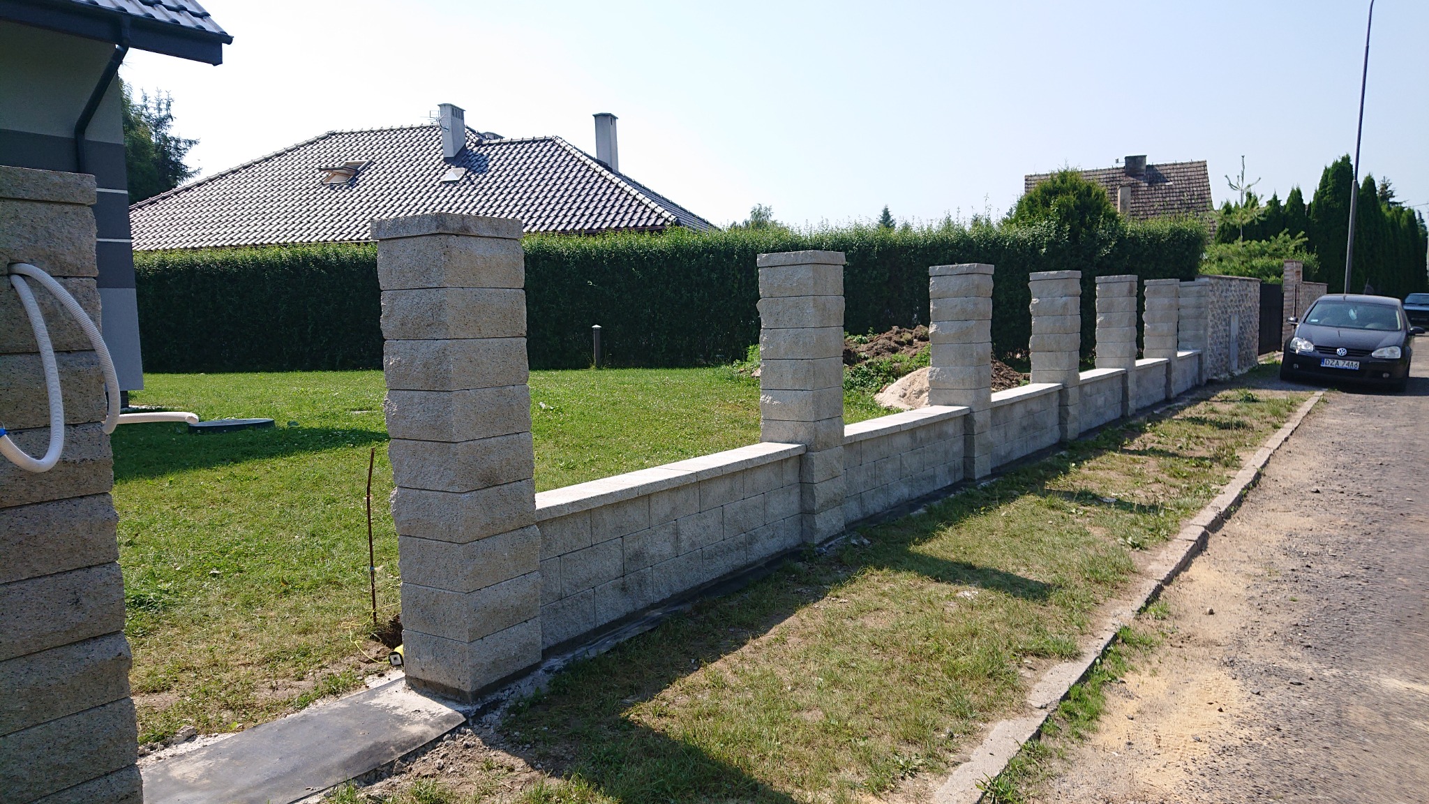 Nowoczesne ogrodzenie modułowe z jasnoszarego betonu architektonicznego, widok z zewnątrz posesji, z fragmentem trawnika i zaparkowanym samochodem w tle.