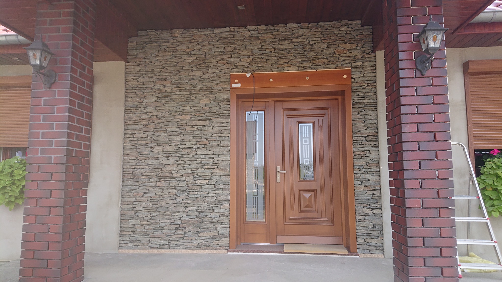 Eleganckie, drewniane drzwi wejściowe z przeszkleniami, otoczone kamienną elewacją i ceglanymi filarami z lampami, widoczne z poziomu gruntu.