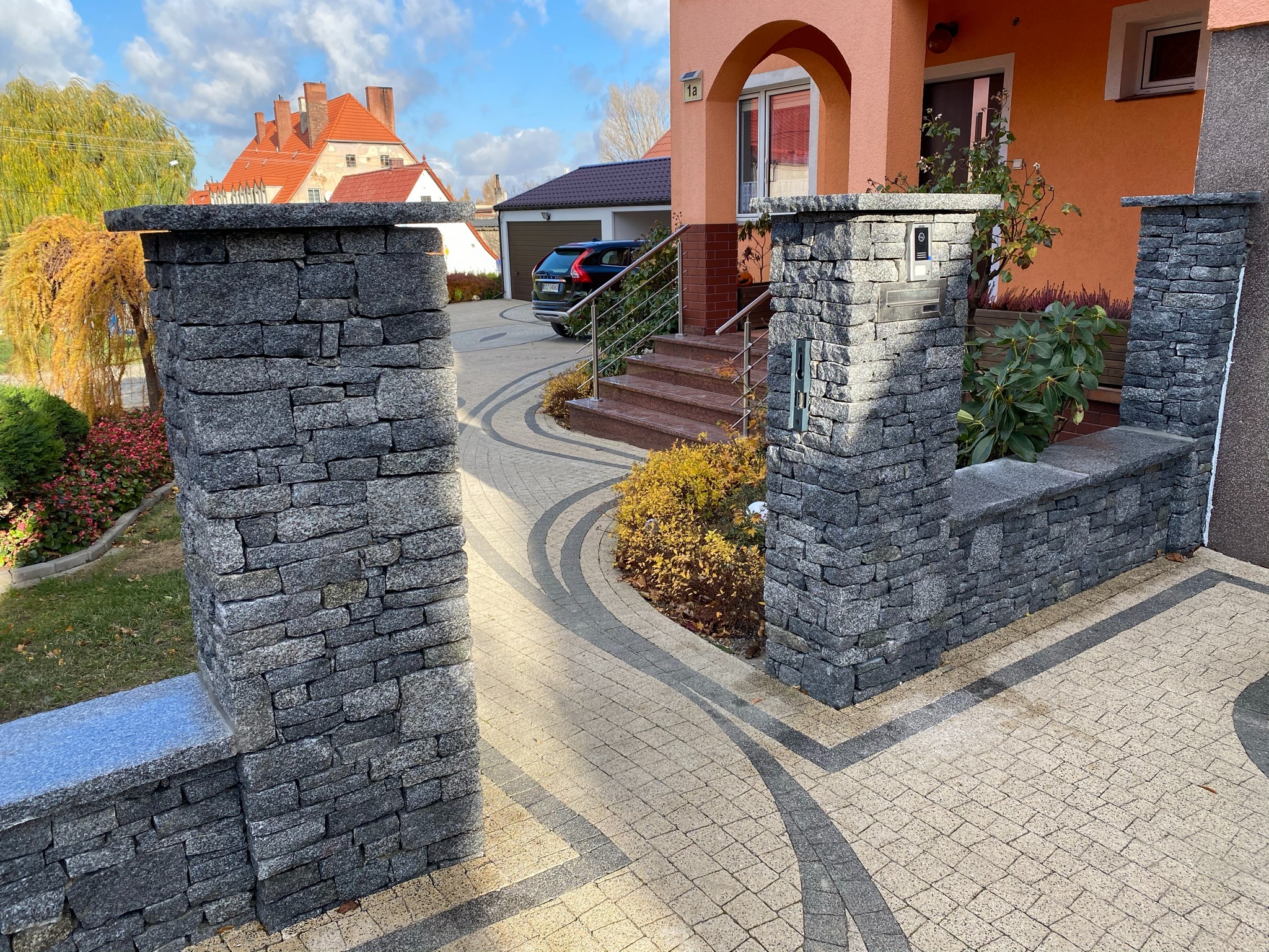 Murowane słupki z szarego kamienia tworzące wejście na posesję z ozdobną kostką brukową, widoczne schody do domu i zaparkowany samochód.