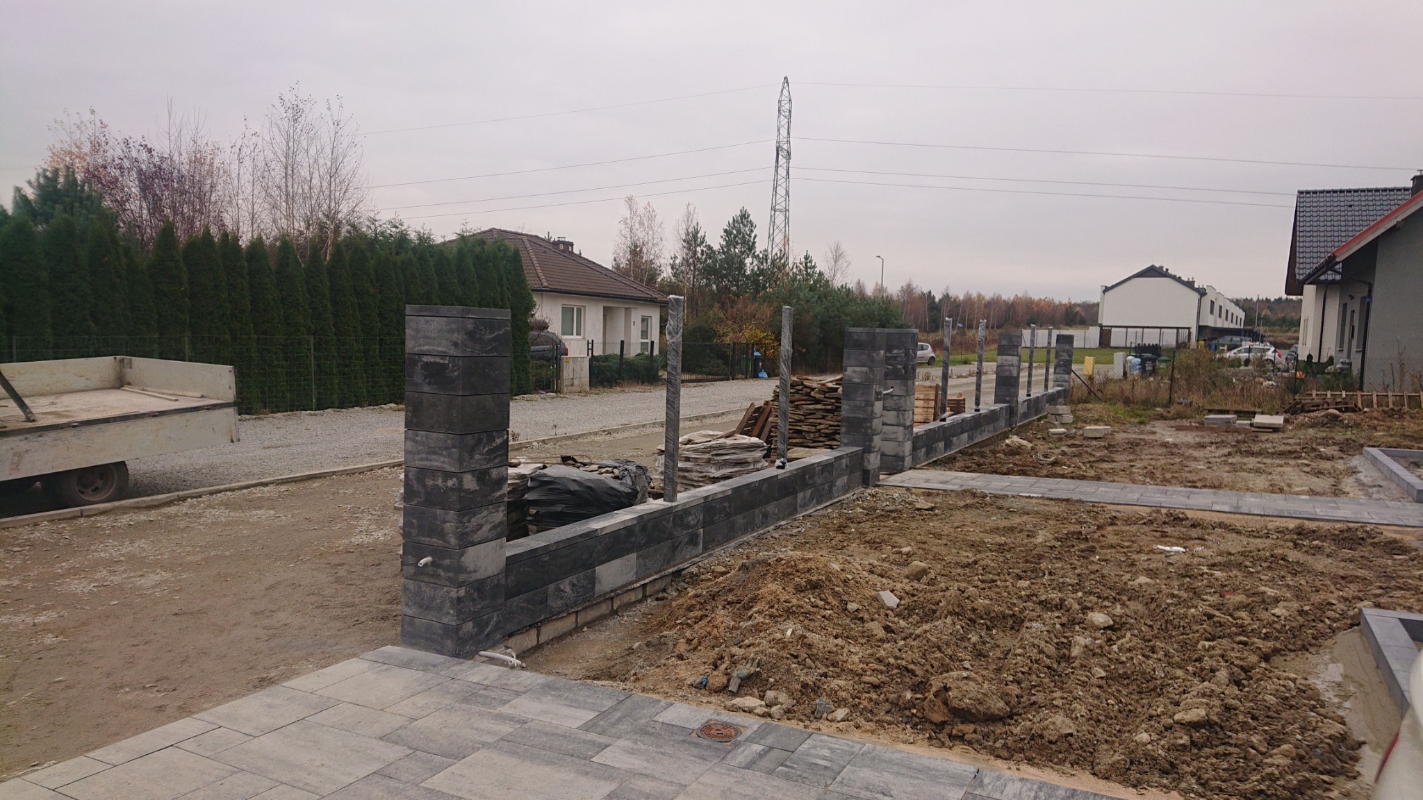 Budowa ogrodzenia z szarej kostki betonowej; widoczne słupki i podmurówka w trakcie montażu na tle domów i pochmurnego nieba.