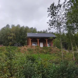 Niewielki drewniany dom z ciemnym dachem na wzgórzu, otoczony zielenią i drzewami w pochmurny dzień. Widoczne elementy konstrukcyjne z drewna i białe drzwi wejściowe.