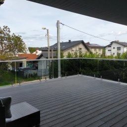 Zenon Więcławek - Nowoczesny taras z desek kompozytowych w kolorze grafitowym, zabezpieczony szklaną balustradą na metalowych słupkach, z widokiem na domy jednorodzinne i zieleń.