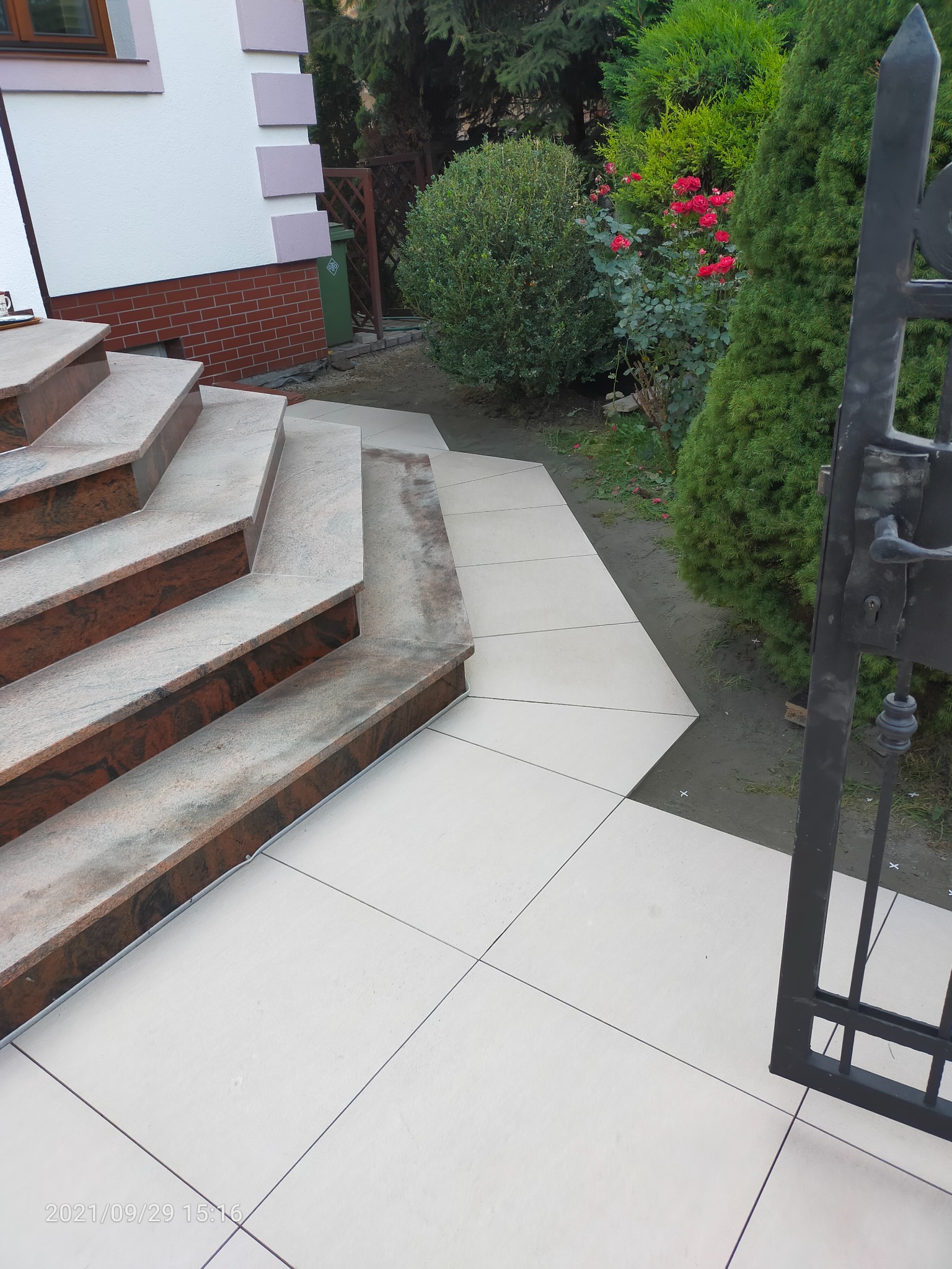 Wyłożone płytkami schody wejściowe z brązowymi akcentami i jasna ścieżka prowadząca od furtki do domu, otoczone zielenią.