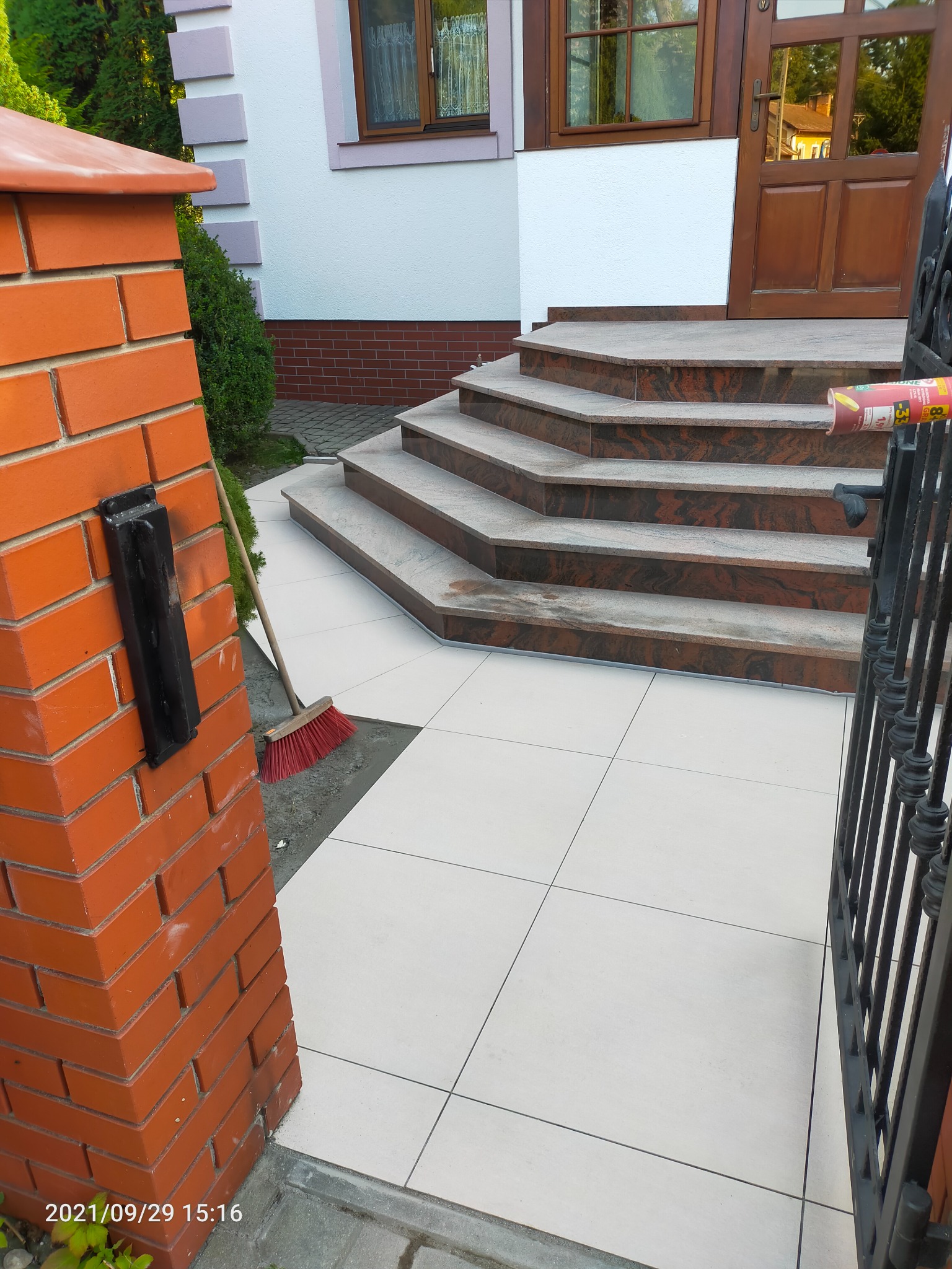 Ułożone białe płytki chodnikowe przed domem z ceglanym słupkiem i schodami wyłożonymi brązowym kamieniem, widoczna szczotka do zamiatania i tuba kleju.
