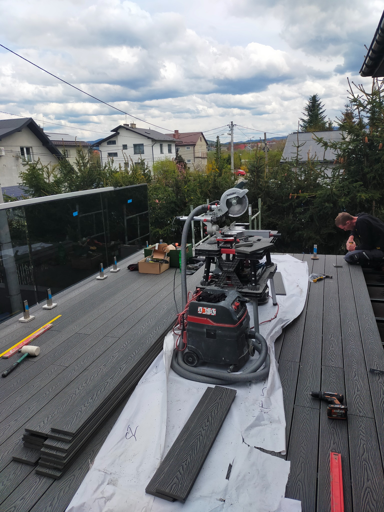 Montaż desek kompozytowych na tarasie z częściowo zamontowaną szklaną balustradą, widoczne narzędzia pracy: piła, odkurzacz przemysłowy, poziomica, w tle zabudowa jednorodzinna.