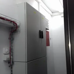 Szara rozdzielnica elektryczna z przewodami w korycie kablowym, widoczne dwa pokrętła regulacyjne i czerwona szafka z wyświetlaczem.