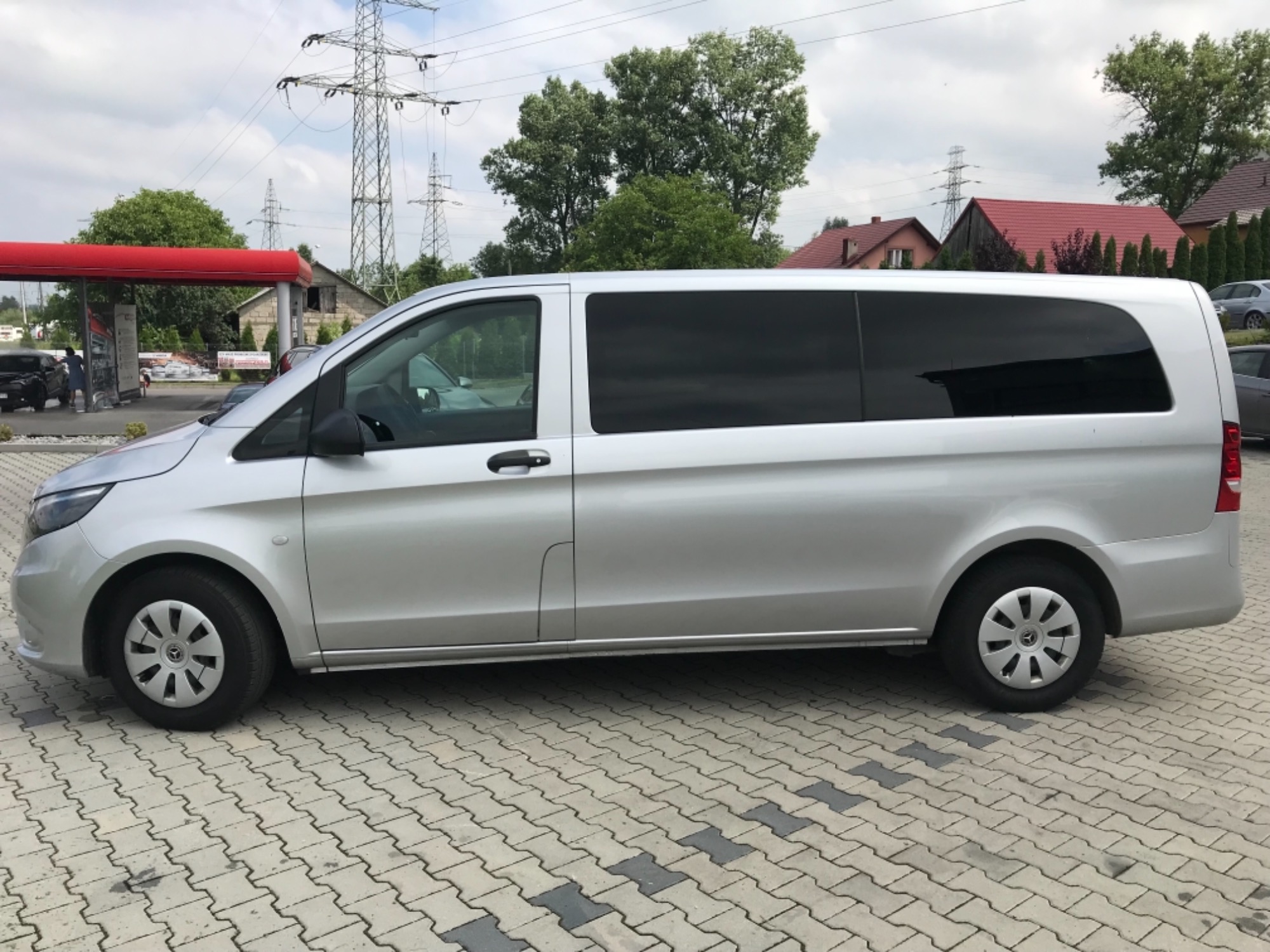 Srebrny van Mercedes Vito z przyciemnionymi szybami zaparkowany na kostce brukowej, widok z boku, w tle słupy wysokiego napięcia.