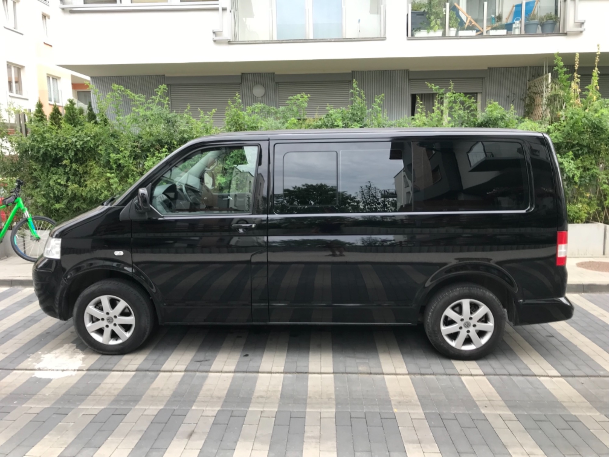 Czarny van Volkswagen Transporter zaparkowany na chodniku z szaro-białych pasów, budynek mieszkalny w tle, rower oparty o krzew.