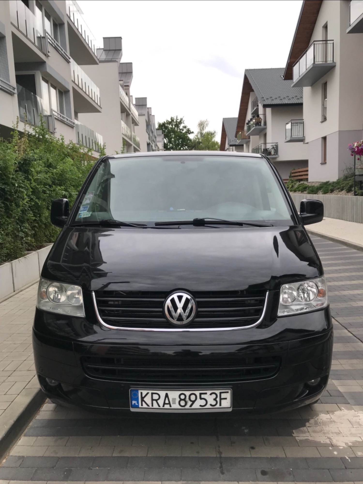 Czarny Volkswagen Transporter T5 z polską tablicą rejestracyjną KRA-8953F, zaparkowany na chodniku z kostki brukowej przed budynkami mieszkalnymi.