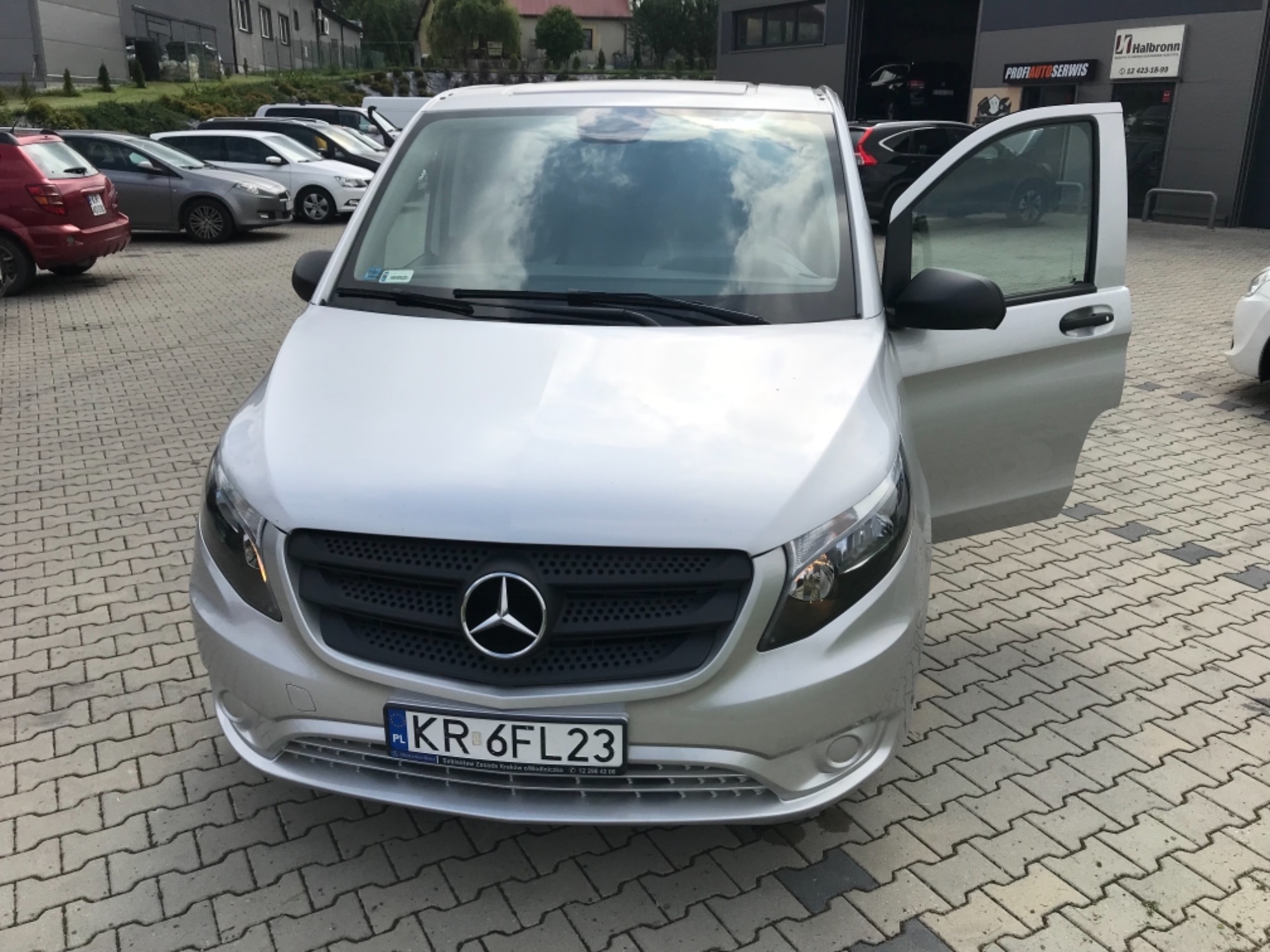 Srebrny Mercedes Vito z otwartymi drzwiami pasażera, zaparkowany na brukowanym placu z innymi samochodami w tle.