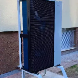 Zewnętrzna jednostka pompy ciepła Hewalex na betonowej podstawie, widoczna aluminiowa konstrukcja wsporcza i fragment elewacji budynku.