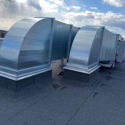 MW-KLIM - System kanałów wentylacyjnych z ocynkowanej blachy na dachu budynku, zabezpieczony odgromnik, widok na miasto w tle pod błękitnym niebem