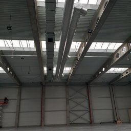 MW-KLIM - Wnętrze hali przemysłowej z widoczną instalacją wentylacyjną pod sufitem, pracownik na podnośniku koszowym w lewym dolnym rogu.