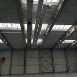 Wnętrze hali przemysłowej z widoczną instalacją wentylacyjną pod sufitem, pracownik na podnośniku koszowym w lewym dolnym rogu.