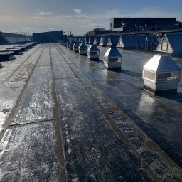 MW-KLIM - Rząd metalowych wywietrzników na pokrytym papą dachu budynku przemysłowego w słoneczny dzień.
