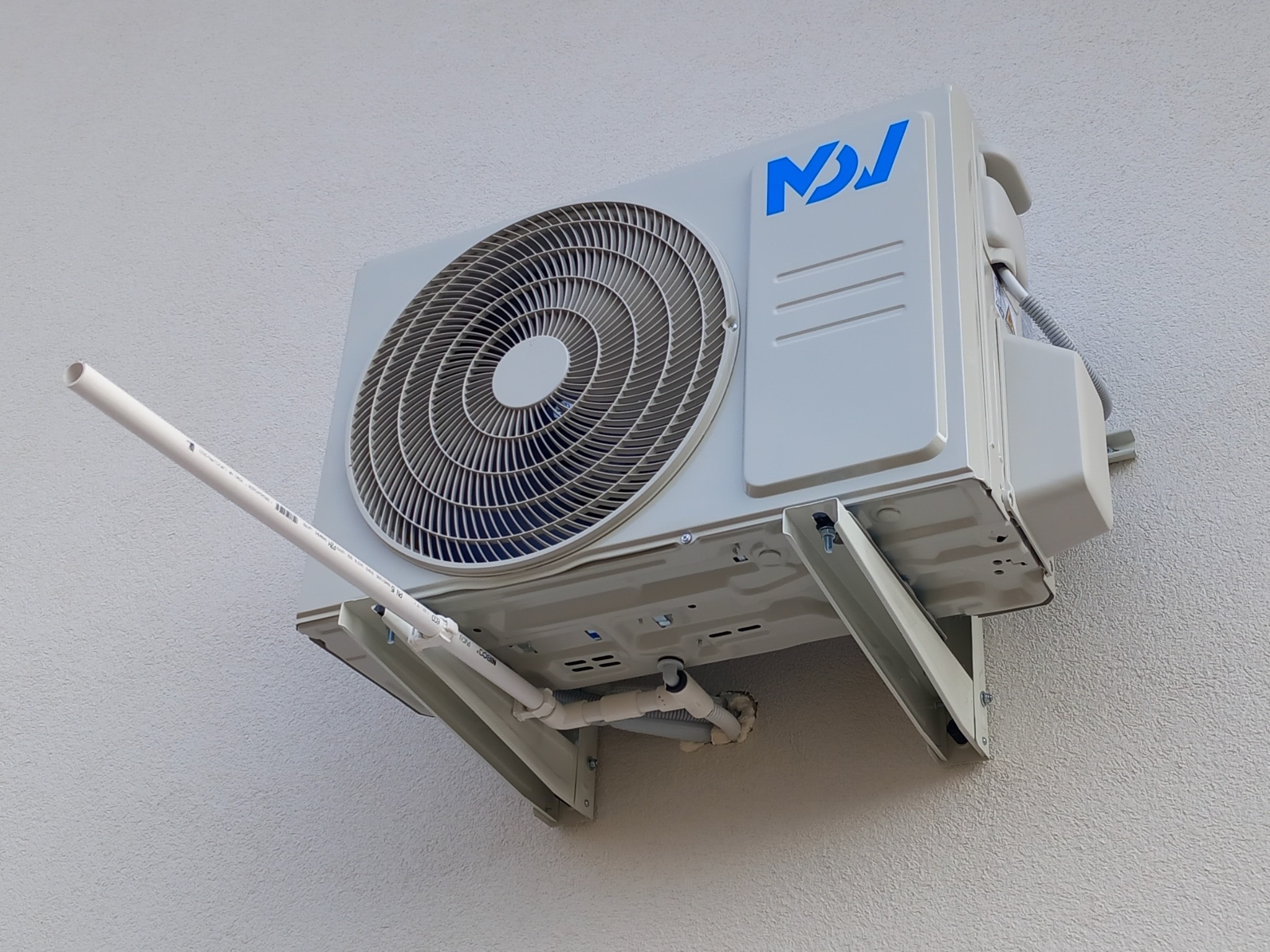 Zewnętrzna jednostka klimatyzacji z widocznym niebieskim logo, zamontowana na ścianie budynku z widocznymi rurami odprowadzającymi skropliny.
